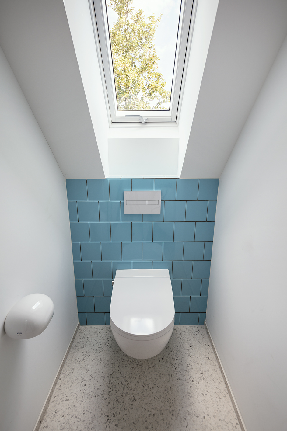 Weißes WC mit Drückerplatte auf blauen Fliesen, Papierrollenhalter und Fenster auf schrägem Dach