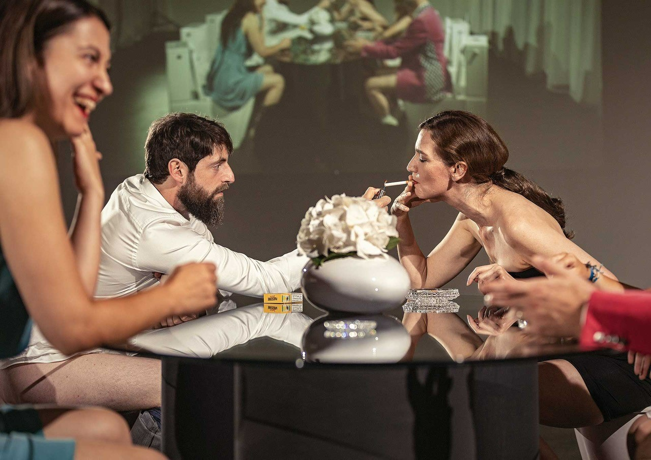 Zwei Frauen und ein Mann sitzen bei einem Tisch und unterhalten sich, eine Frau raucht eine Zigarette