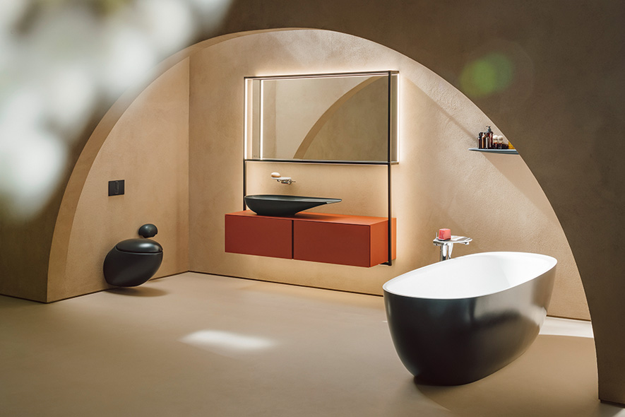 Badezimmer mit erd-tönigen Wänden, schwarz-weiße freistehende Badewanne, schwarze Keramikwaschtisch auf terracotta farbenem Möbel, darüber beleuchteter Spiegel, links schwarzes WC