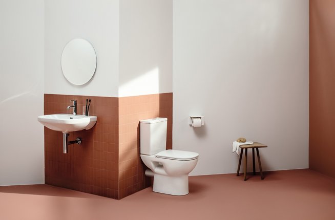 Badezimmer mit orange-farbenen Wand- und Bodenfliesen, an der Wand hängt ein weißer Waschtisch mit Armatur und Spiegel, daneben ein weißes Stand-WC 